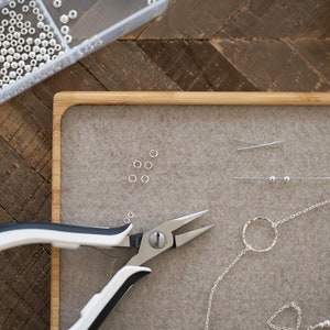 May include: A close-up of a jewelry making workspace with a tray of silver beads, a pair of silver pliers, a silver chain, and a silver ring.