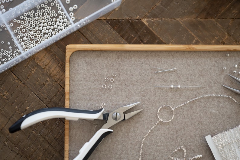 May include: A close-up of a jewelry making workspace with a tray of silver beads, a pair of silver pliers, a silver chain, and a silver ring.