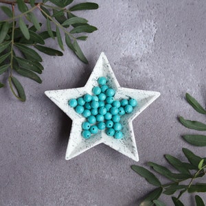 May include: A white star-shaped dish with a speckled finish holds a collection of turquoise blue round beads.