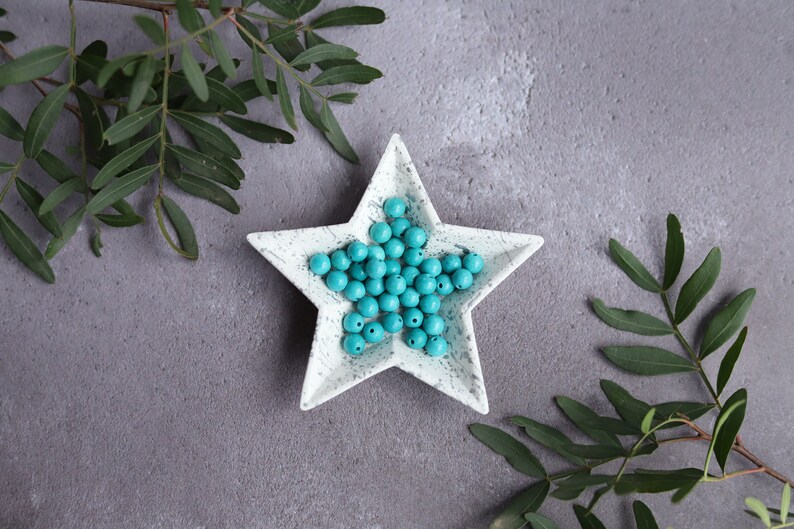 May include: A white star-shaped dish with a speckled finish holds a collection of turquoise blue round beads.