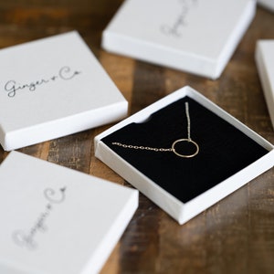 May include: A delicate gold circle necklace in a white box with the text "Ginger & Co" on the lid. The necklace is displayed on a black velvet cushion.