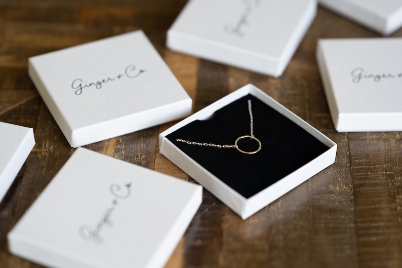 May include: A delicate gold circle necklace in a white box with the text "Ginger & Co" on the lid. The necklace is displayed on a black velvet cushion.