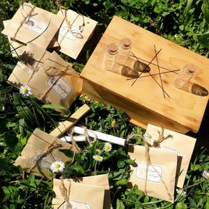Peut inclure: Une boîte en bois avec un motif d'étoile et quatre petites bouteilles en verre. La boîte est entourée d'enveloppes en papier brun avec de la ficelle et des étiquettes qui disent "Charms of the Wild".