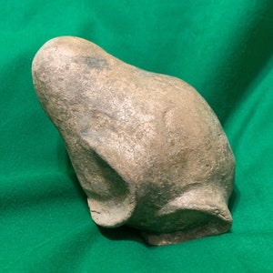 以下が含まれることがあります： 風化した、明るい茶色の彫刻されたオブジェクトで、丸みを帯びた上部と基部があります。オブジェクトは粗い質感で、石または粘土でできているようです。形状は有機的で抽象的で、おそらく様式化された動物または人物です。