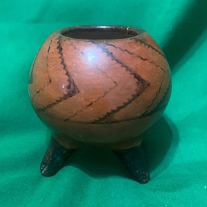 May include: A small, round, hand-painted ceramic pot with three dark legs. The pot is brown with dark zigzag patterns and a dark interior. The pot is sitting on a green surface.