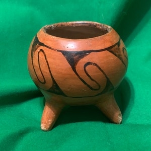 May include: A small, round, terracotta-colored ceramic pot with three short legs. The pot features a dark, swirling design around its exterior. The interior is dark, and the pot sits on a green surface.