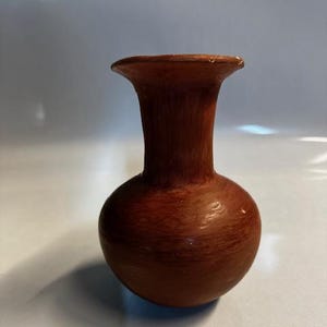 May include: A small, handcrafted, brown vase with a rounded body and a flared rim. The vase has a smooth, matte finish and appears to be made of ceramic or clay. The vase is likely used for decorative purposes or to hold small flowers.