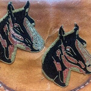 May include: Two black horse head appliques with gold trim, sequined muzzles, and red accents. The appliques are on a brown leather surface. The horse heads are facing left and are detailed with black beads.