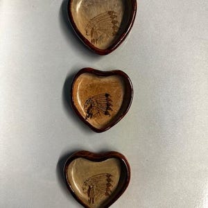 May include: Three heart-shaped wooden dishes with a dark brown rim and a light brown interior. Each dish features a Native American chief design. The dishes are arranged vertically against a light gray background.