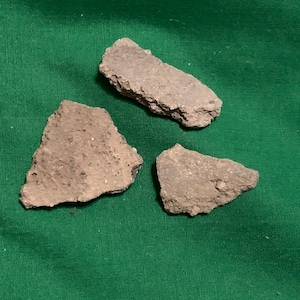 Puede incluir: Tres fragmentos de cerámica de color gris-marrón de forma irregular descansan sobre una tela verde. Los fragmentos tienen bordes ásperos y texturizados y tamaños variables, lo que sugiere que son restos de un objeto más grande.