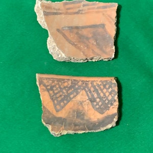 Puede incluir: Dos fragmentos de cerámica antigua con diseños geométricos descoloridos en naranja y negro. Los fragmentos de cerámica tienen bordes ásperos e irregulares y se asientan sobre un fondo verde. Los diseños incluyen líneas y un patrón de entramado.