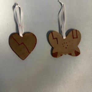 May include: Two handmade ornaments: a heart and a butterfly. Both are brown with red accents and white ribbon hangers. The heart has a broken line design, and the butterfly has a symmetrical pattern.