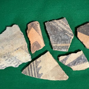 May include: A collection of ancient pottery shards with geometric designs. The fragments display a variety of colors, including tan, black, and orange, and are arranged on a green surface. The pottery pieces have unique shapes and patterns.