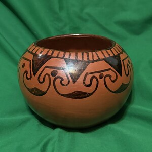 May include: A round, terracotta-colored ceramic bowl with a dark brown rim and decorative black geometric patterns. The bowl has a smooth surface and a wide opening, suitable for various uses.