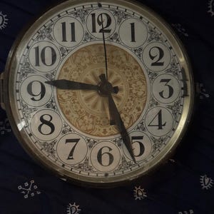 Può includere: Un orologio rotondo in stile antico con quadrante color crema e numeri neri. Le lancette dell'orologio indicano circa le 9:27. Il quadrante presenta un motivo floreale decorativo in oro e nero, racchiuso in una cornice metallica. Un oggetto di arredamento vintage.
