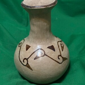 May include: A beige ceramic vase with a rounded body and a narrow neck. The vase features dark brown, stylized geometric designs. The rim is accented with a darker brown band. The vase is displayed against a green background.