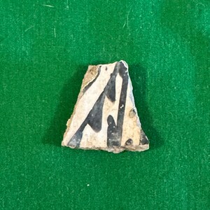 May include: A fragment of pottery with a cream-colored base and black painted design. The design features abstract shapes and lines. The edges of the pottery are rough and uneven, suggesting it is an antique or relic.