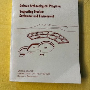 May include: A vintage book cover with the title "Dolores Archaeological Program: Supporting Studies: Settlement and Environment." The cover features a line drawing of a landscape with a structure and is published by the U.S. Department of the Interior.