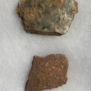 以下が含まれることがあります： 形が不規則な2つの岩石標本。 上部の標本は、わずかに反射する表面を持つ、層状の茶色と灰色の石です。 下部の標本は、より明るい色の粒子が散りばめられた、ざらざらした質感の外観の赤茶色の石です。