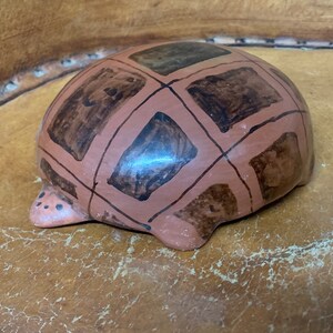 May include: A ceramic turtle figurine with a reddish-brown shell featuring a grid pattern of dark brown squares. The turtle's head and feet are a lighter shade of brown. The figurine is a decorative item.