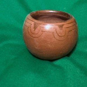 May include: A small, round, brown ceramic bowl with a decorative etched pattern around the top. The bowl has a smooth, unglazed surface and a wide opening. The bowl is likely handmade and suitable for holding small items or as a decorative piece.