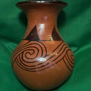 May include: A terracotta-colored vase with a flared rim and a rounded body. The vase features black painted designs, including two triangles near the top and swirling lines across the body. The rim is dark brown.