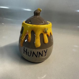 May include: A ceramic honey pot with a brown lid and a yellow honey-like design dripping down the sides. The pot is grey with the word "HUNNY" in black. A small bee is on the side of the pot.