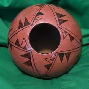 May include: A round, terracotta-colored ceramic vessel with a large central opening. The surface is decorated with a repeating pattern of black triangular shapes and lines. The vessel sits on a green surface.