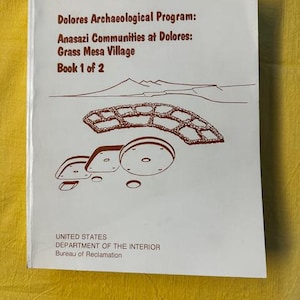May include: A white book cover with the title "Dolores Archaeological Program: Anasazi Communities at Dolores: Grass Mesa Village Book 1 of 2." The cover features a brown illustration of a structure and mountains.