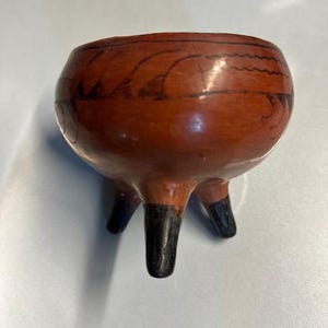May include: A small, round, terracotta-colored ceramic bowl with three black legs. The bowl has a dark, wavy design around the rim. The legs are painted black at the bottom.