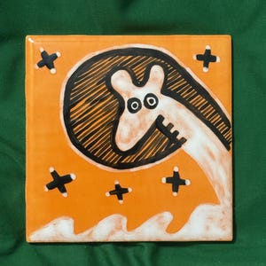 May include: Square orange ceramic tile featuring a whimsical cartoon giraffe-like creature with a black and white design. The tile has black cross shapes and white wave-like details. The background is a solid green.