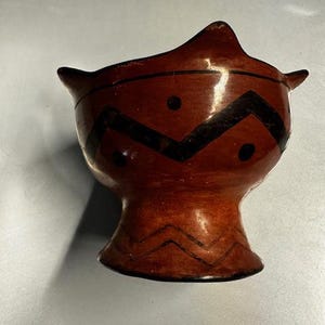 May include: A brown ceramic bowl with a zig-zag and dot pattern in black. The bowl has a flared rim with two small points and a pedestal base. The bowl is hand-painted with a glossy finish.