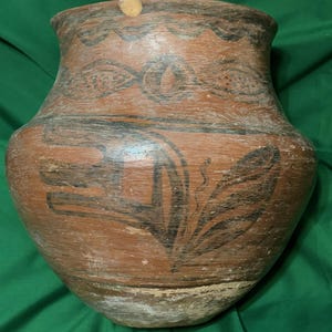 May include: A large, antique terracotta pot with a wide body and narrow neck. The pot features black painted designs, including geometric shapes and floral motifs. The exterior has a weathered, aged appearance, suggesting its historical significance.