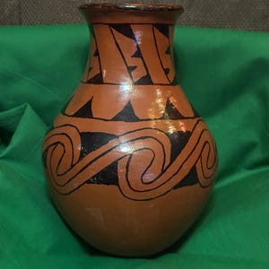 May include: A terracotta-colored vase with black geometric patterns. The vase features a wide body, a narrow neck, and a flared rim. The design includes triangles and wave-like shapes, creating a handcrafted aesthetic. The vase is displayed on a green surface.