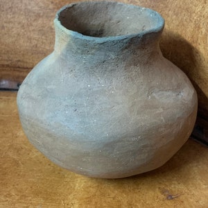 Puede incluir: Una olla de barro hecha a mano, de forma redonda, con un cuerpo ancho y un cuello estrecho. La olla es de color marrón apagado con una textura ligeramente rugosa. El borde de la olla es de un tono marrón más oscuro.