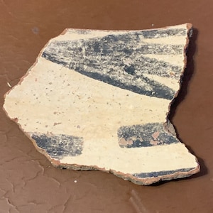 May include: Fragment of an ancient ceramic vessel. The pottery shard features a cream-colored background with black painted geometric designs. The edges show the original reddish-brown clay.