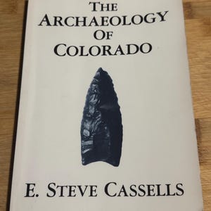 Peut inclure: Un livre à couverture souple intitulé The Archaeology of Colorado par E. Steve Cassells. La couverture blanche présente une illustration centrée, sombre et détaillée d'une pointe de projectile en pierre, avec le titre et l'auteur en noir.