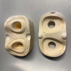 Puede incluir: Dos formas abstractas de cerámica blanquecina, con bordes redondeados y aberturas circulares. Una es rectangular, la otra ovalada. Ambas tienen un aspecto texturizado y envejecido, lo que sugiere un origen vintage o artesanal. Las formas están sobre una superficie gris.