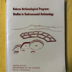 May include: A book cover with the title "Dolores Archaeological Program: Studies in Environmental Archaeology" in maroon text. The cover features line drawings of a landscape and archaeological site. The bottom of the cover reads "UNITED STATES DEPARTMENT OF THE INTERIOR Bureau of Reclamation."