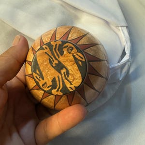 May include: A round, wooden decorative object with a sunburst design. The object features a central circular emblem with a stylized animal figure in black and brown. The sunburst design has red and brown accents.