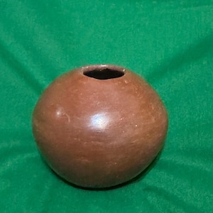May include: A small, round, brown ceramic pot with a dark opening. The pot has a smooth, slightly glossy surface and sits against a green background. The pottery appears handmade.