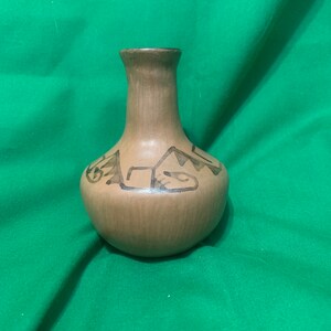 May include: A small, handcrafted vase with a rounded body and a narrow neck. The vase is a muted brown color with darker brown geometric designs. The vase is likely made of ceramic or clay and is suitable for small floral arrangements.