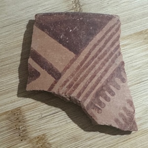 May include: A fragment of ancient pottery with a geometric design. The pottery shard is reddish-brown with a pattern of dark red lines and shapes. The design includes diagonal stripes and angular forms.