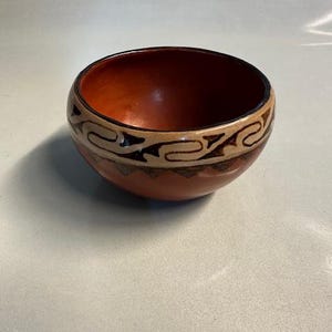 May include: A small, round, hand-painted bowl with a reddish-brown interior and exterior. The rim features a decorative band with a repeating pattern in tan and black. The bowl is likely made of ceramic or clay.