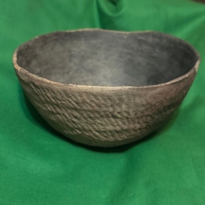 May include: A handmade, textured bowl with a dark gray interior and a rough, brown exterior. The bowl is set against a green background, showcasing its unique, organic form. The bowl's rim is a lighter shade of brown, adding contrast.