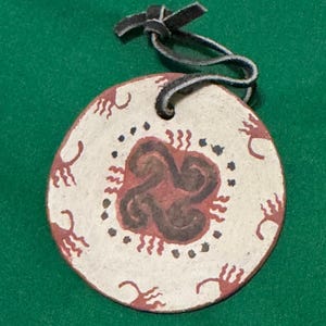 May include: A round, off-white ornament with a dark brown and red design. The ornament has a black leather loop for hanging. The design features a central spiral and surrounding figures.