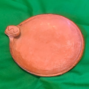 May include: A round, terracotta-colored ceramic plate with a raised edge and a small, decorative handle. The plate has a smooth surface and a rustic, handmade appearance. The plate is set against a green background.