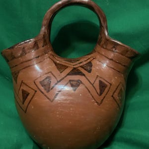 May include: A brown ceramic vase with a handle, featuring geometric patterns in dark brown. The vase has a wide opening and a rounded body, perfect for decorative use.