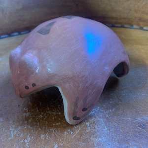 May include: A handmade, terracotta-colored ceramic helmet with a rounded top and a curved, open base. The helmet has dark brown accents and two small, dark dots near the base. The surface has a slightly rough texture.