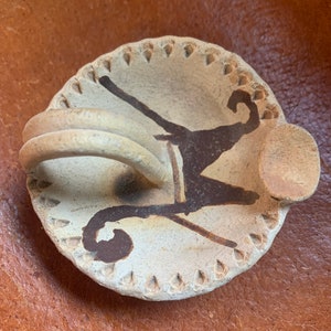 May include: A small, off-white ceramic dish with a raised handle and a circular base. The dish features a dark brown abstract design and a decorative edge with small, triangular cutouts. The handle is a coiled design.